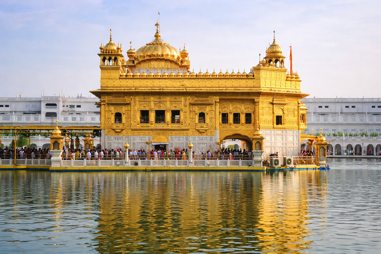 A side-profile view of the Golden Temple in Amritsar, India, showing the gilded gold structure and its reflection in the surrounding Amrit Sarovar (sacred pool) under a bright daylight sky.