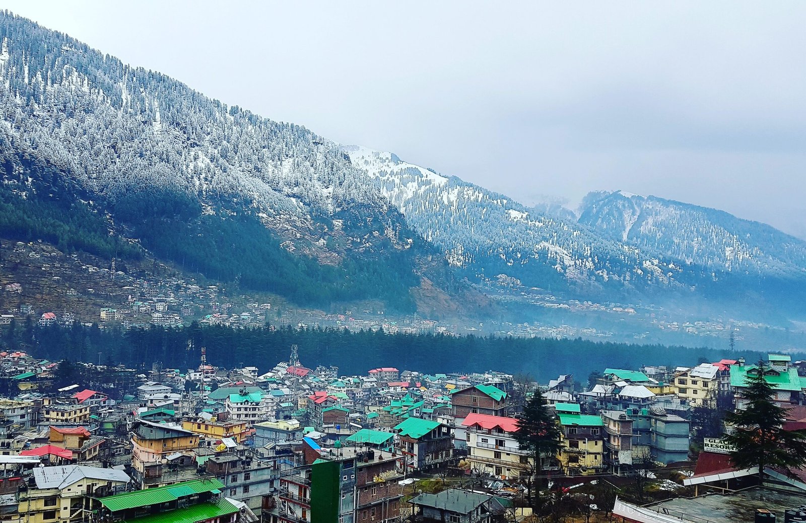 A high-angle panoramic view of Manali town showing colorful rooftops and buildings clustered in the valley, with massive snow-dusted pine forests and Himalayan mountains in the background.