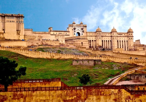 A wide daytime shot of the historic Amer Fort in Jaipur, Rajasthan, showcasing its massive yellow sandstone walls, intricate ramparts, and hilltop position against a blue sky.